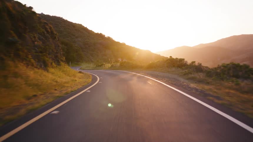 A first-person view of a scenic drive along a winding mountain road reveals the warm, glowing sunset