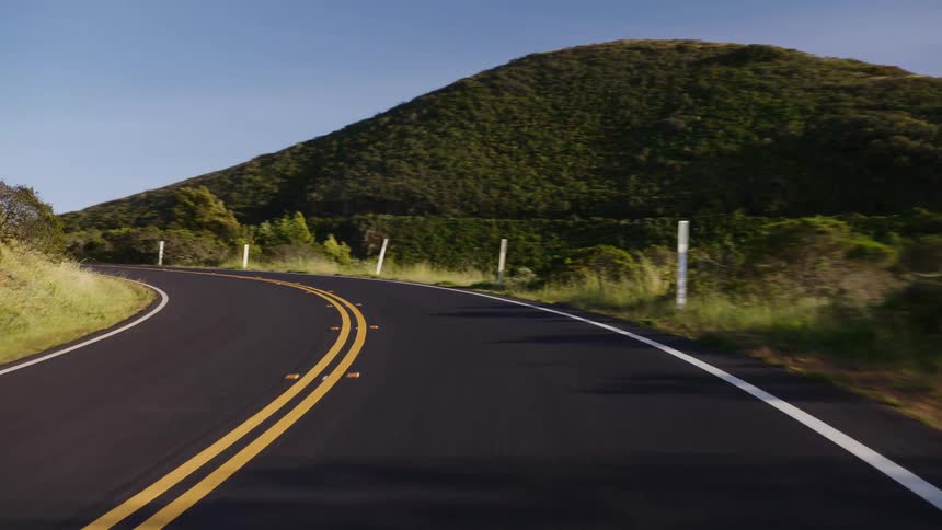 A smooth point-of-view drive follows the curve of a scenic, winding road through lush green hills on a bright, sunny day
