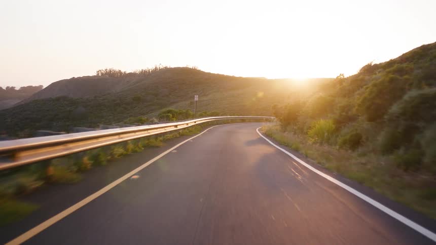 A serene point-of-view drive along a winding mountain road as the golden sun sets over the hills