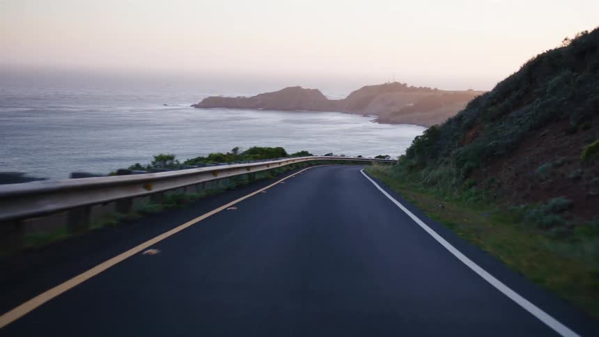 A serene point-of-view drive along a winding coastal highway with the ocean visible as the sun begins to set