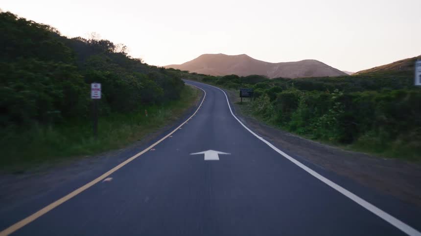 A serene point-of-view drive along a winding rural road through green hills as the sun sets over distant mountains