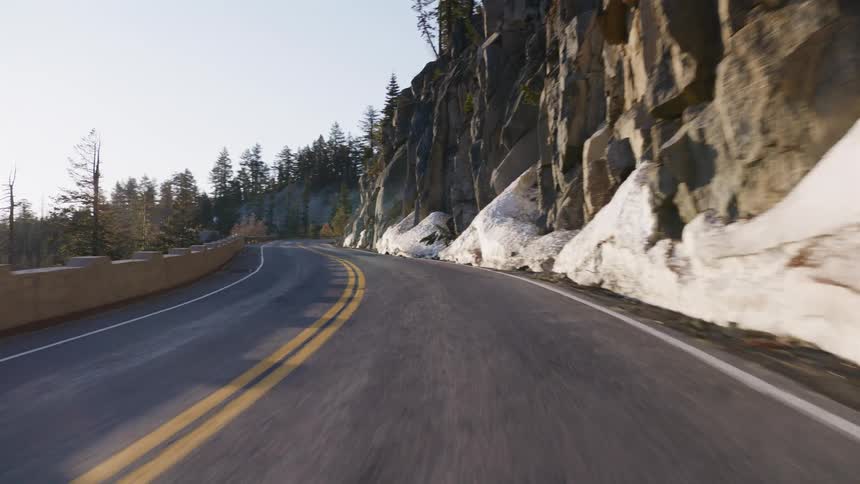 A smooth point-of-view drive along a winding mountain road with snow-covered rocks and pine trees in the late afternoon sun