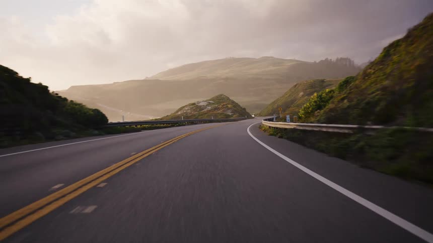 A point-of-view shot driving along a scenic coastal highway through rolling hills at sunset