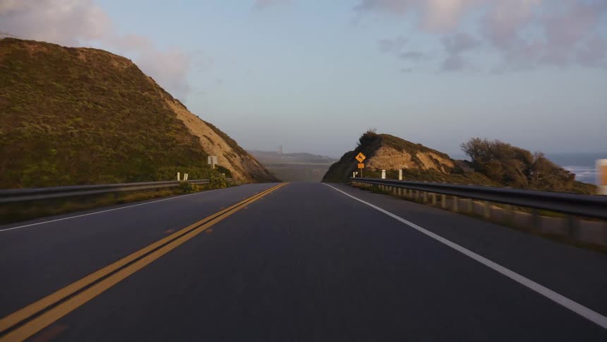 A first-person view driving along a winding coastal highway flanked by sandy bluffs at dawn