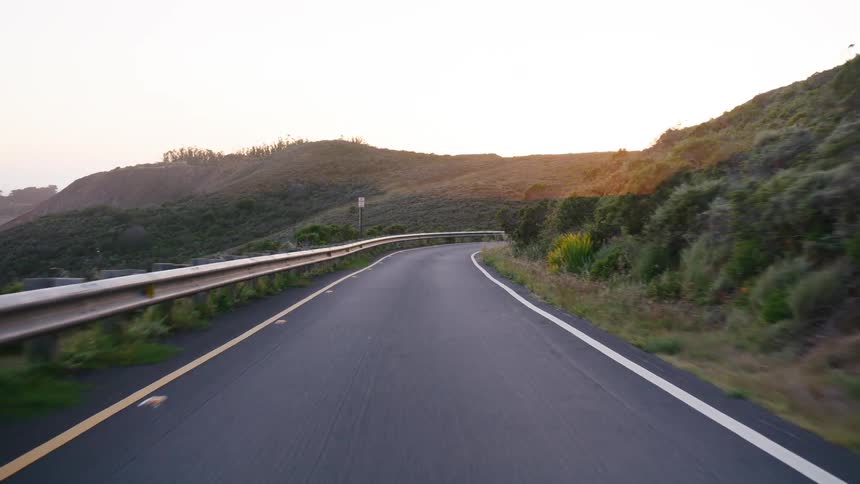 A smooth point-of-view drive along a winding asphalt road through rolling green hills at sunset