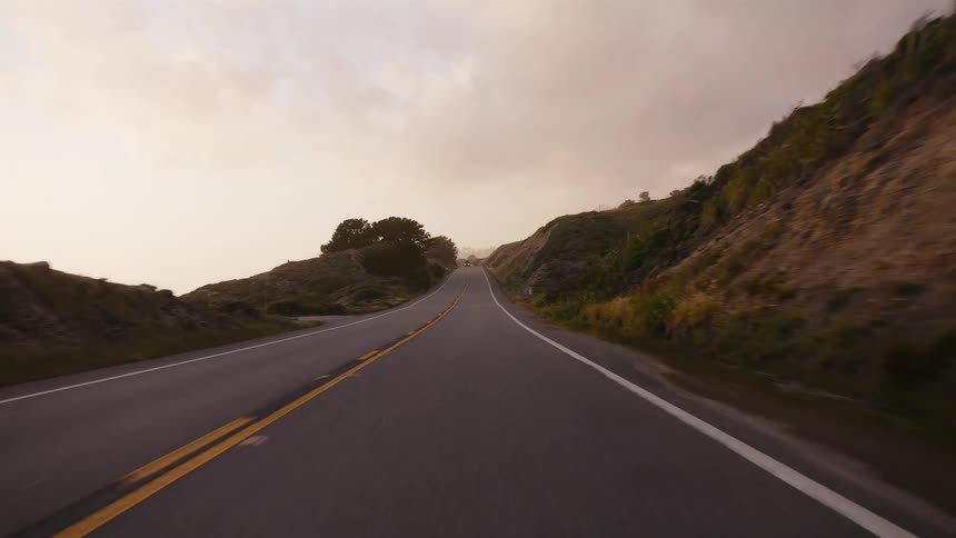A first-person point of view driving on a scenic highway through rolling hills during a hazy sunset