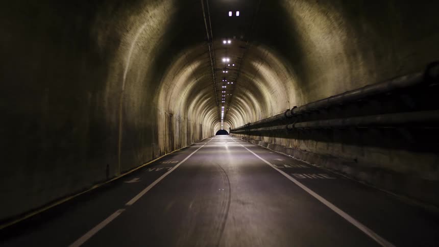 POV shot driving through a long, illuminated concrete road tunnel and emerging onto a winding road at twilight