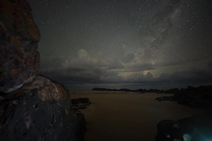 A stunning night timelapse captures clouds parting to reveal the starry Milky Way over a rocky beach as a distant lightning storm flashes on the horizon