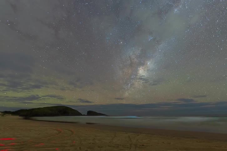 A stunning timelapse captures the Milky Way and faint green airglow moving across the starry night sky over a serene ocean beach