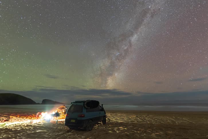 A time-lapse of people enjoying a campfire on the beach under a starry night sky with the Milky Way galaxy and Aurora Australis visible