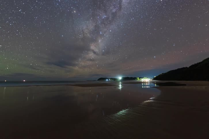 A stunning night-lapse captures the Milky Way galaxy moving across the sky above a tranquil beach, with stars reflecting on the wet sand