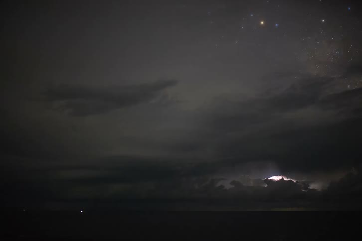 A powerful electrical storm with frequent lightning flashes illuminates the clouds and night sky over the ocean