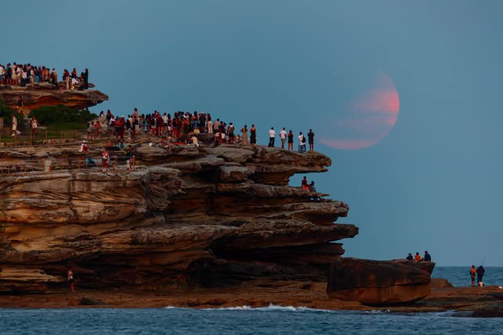 A time-lapse shows a crowd of people gathered on a rocky coastline at dusk to watch a dramatic red moon rise over the ocean