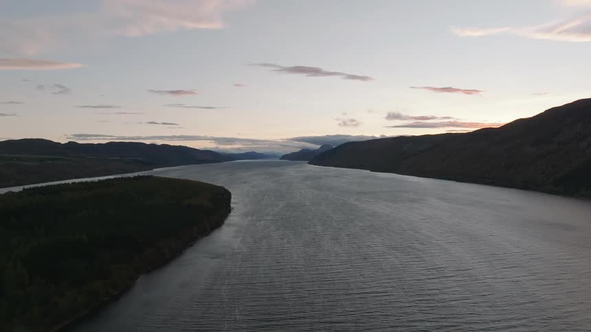 A tranquil aerial view captures the vast expanse of Loch Ness at dusk, nestled between the dark, rolling hills of the Scottish Highlands