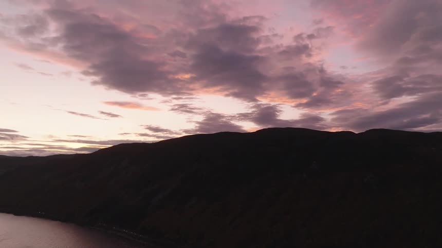 A slow aerial ascent reveals a spectacular purple and pink sky at dusk over a fjord and mountains
