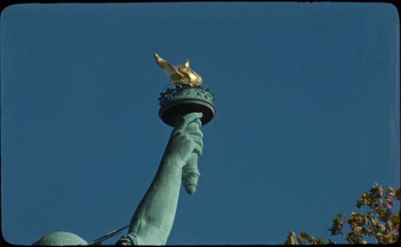 An archival 16mm film style shot shows a low-angle view of the Statue of Liberty's torch against a clear blue sky