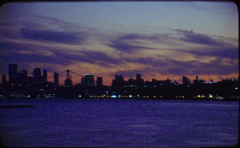 A vintage 16mm film captures a slow pan across the New York City skyline at dusk, with buildings silhouetted against a colorful sunset sky