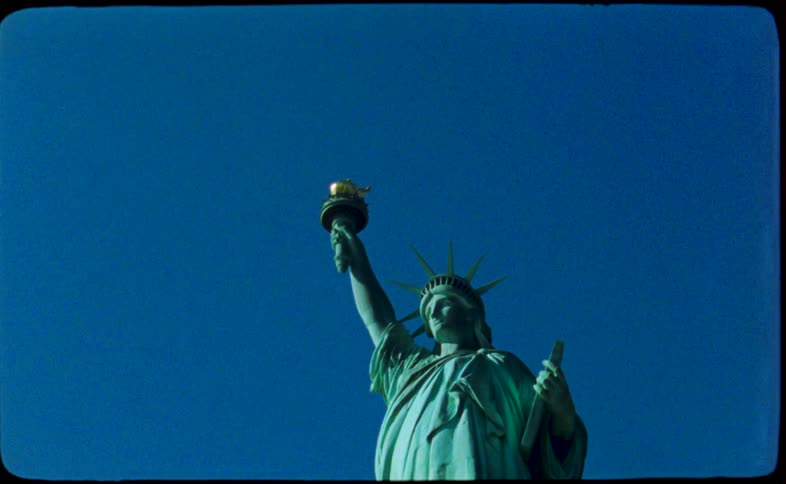 An iconic low-angle tilt shot on vintage film reveals the Statue of Liberty standing tall against a vibrant blue sky