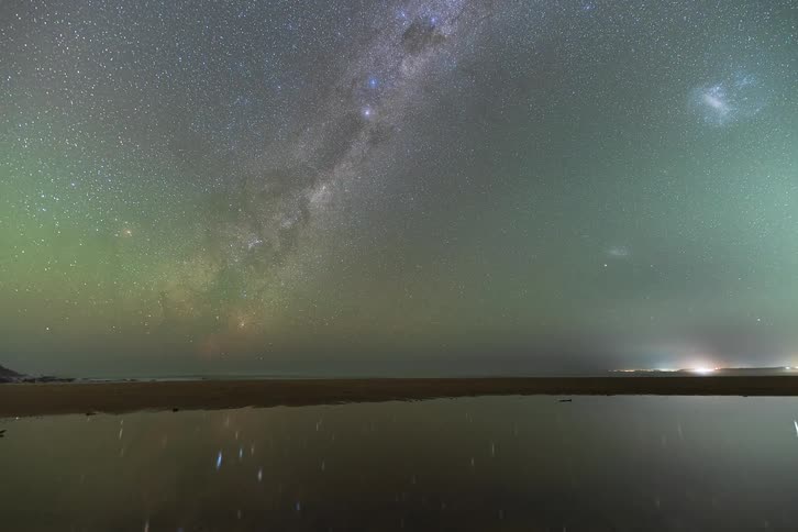Breathtaking time-lapse of the Milky Way galaxy and a vibrant green airglow reflecting in calm waters on a beach before a vivid moonrise