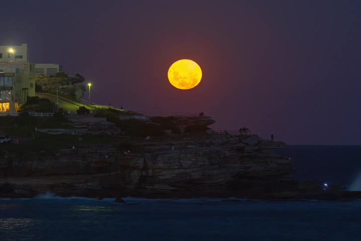 An awe-inspiring timelapse captures a giant orange supermoon rising into the night sky over a rocky seaside cliff as waves crash below