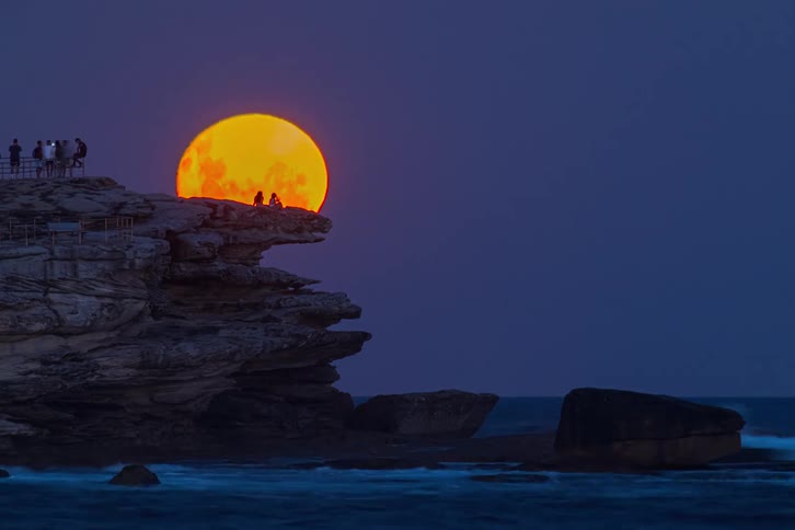 A stunning timelapse captures a vibrant orange supermoon rising over the ocean as people watch from the rocky cliffs