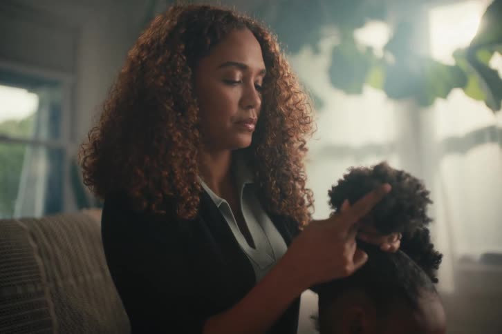 A mother lovingly styles her young daughter's hair while the girl sits on the floor and plays with her doll