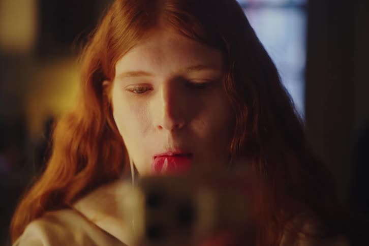 Close-up of a young redhead blowing a pink bubble gum bubble that pops while she watches her smartphone at night