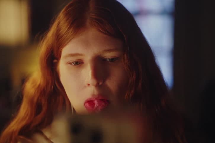 Close-up shot of a young woman with long red hair blowing a bubble with gum while looking at her smartphone at night