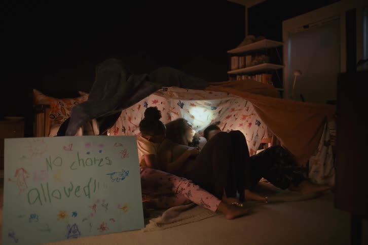 A mother tells a story with a flashlight to her two children as they cuddle together inside a homemade blanket fort at night