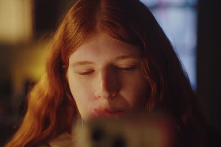 A close-up of a young woman with long red hair watching her phone and blowing a bubble gum in a dimly lit room