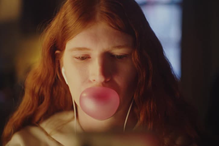 Close-up slow motion shot of a young redhead woman blowing and popping a pink bubble gum bubble while wearing earphones