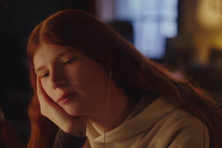 A thoughtful young redhead with earphones rests her head on her hand while listening to music at night