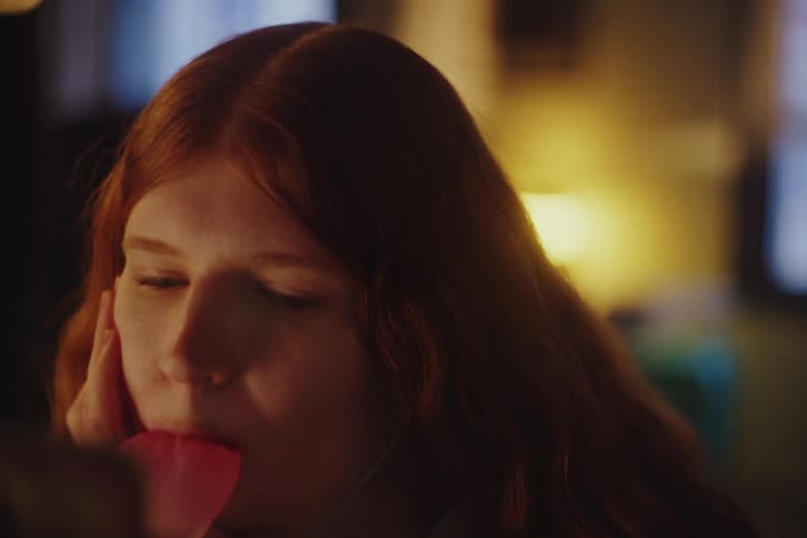 A contemplative young woman with long red hair slowly blows a pink bubblegum bubble in a dimly lit room
