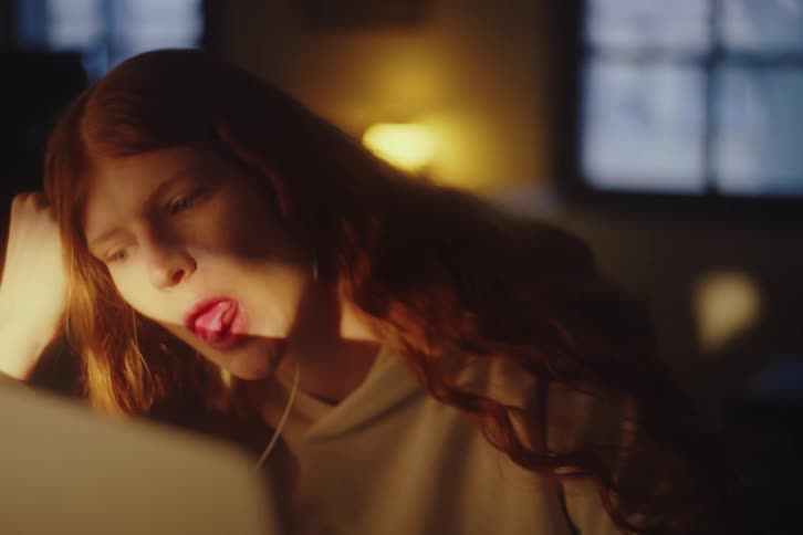 Close-up slow motion shot of a bored young woman with red hair blowing and popping a pink bubble in a dimly lit room
