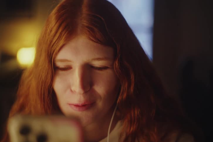 A happy young woman with long red hair has a video call on her smartphone in a dimly lit room