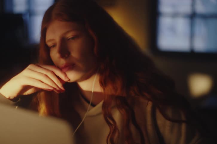 A stressed young woman with long red hair works late at night on her laptop, concentrating in a dimly lit room