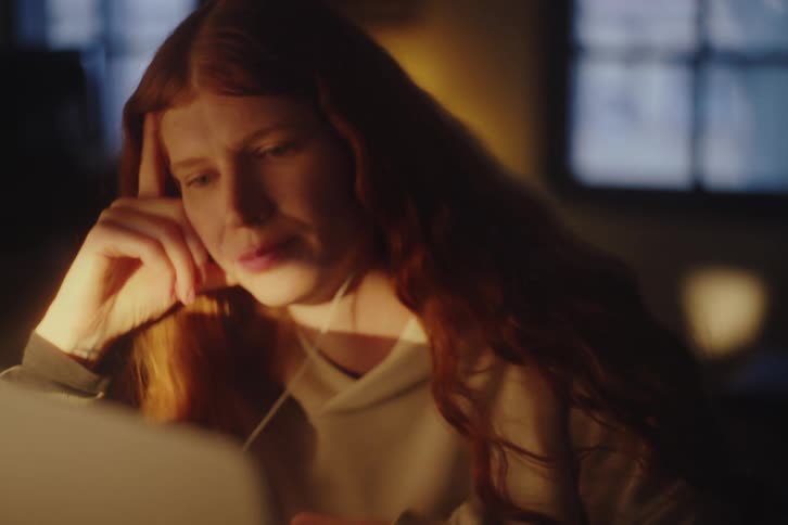 A focused redhead works intently on her laptop in a dimly lit room, becoming stressed and rubbing her forehead in fatigue