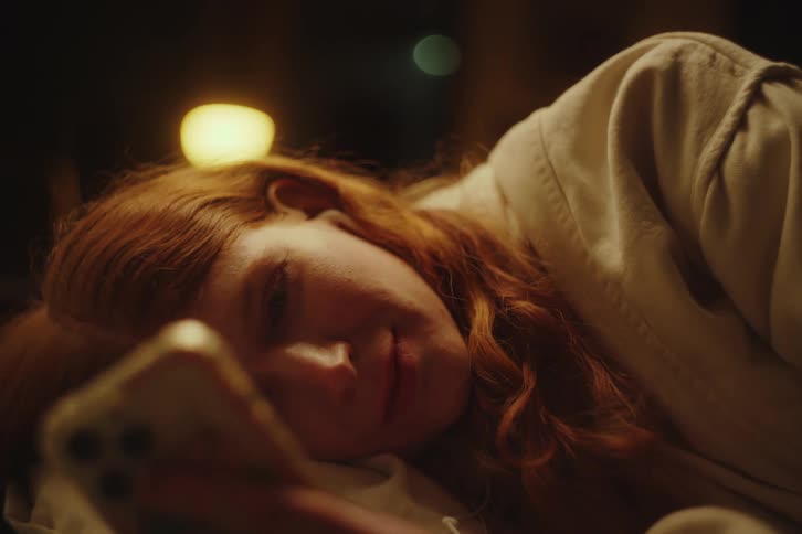 Intimate close-up of a happy young woman with red hair lying in bed at night, smiling as she scrolls on her smartphone