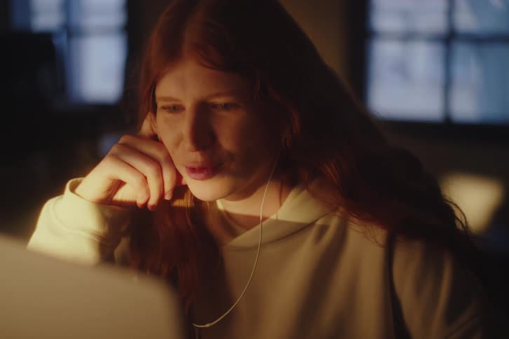 A young woman with long red hair has an engaging video call on her laptop late at night in a dark room
