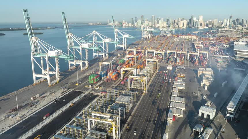 Static aerial view of a busy industrial shipping port with cranes and containers, as trucks and machinery operate with a city skyline in the distance