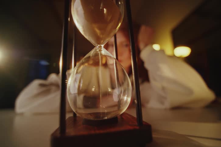 In a low-lit, moody setting, a person's hand turns over an hourglass as they watch the sand fall, representing the passage of time