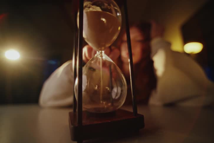 A close-up shot shows a hand flipping an hourglass on a table, resetting the flow of sand in a moody, dimly lit room