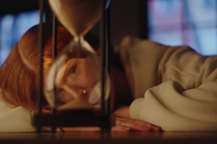 A melancholic redhead woman rests her head on a table, watching time slip away through an hourglass in a dimly lit room