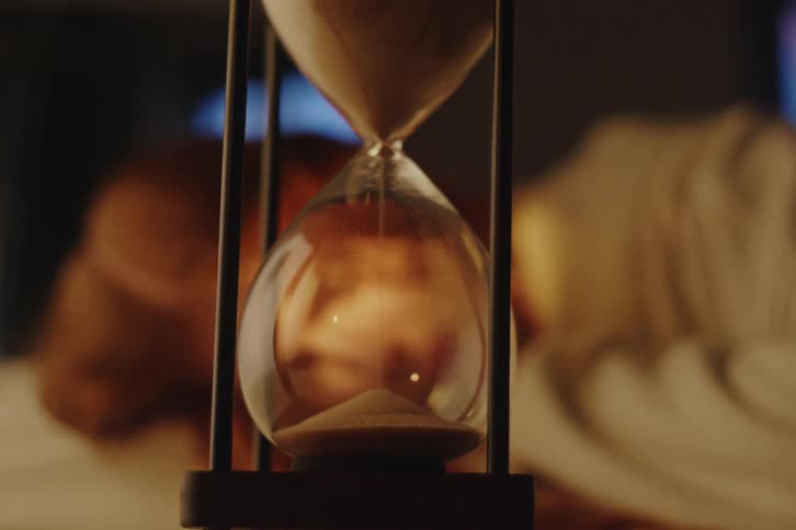 Cinematic close-up of an hourglass with sand slowly falling as a person sleeps in a dark, warm-lit room