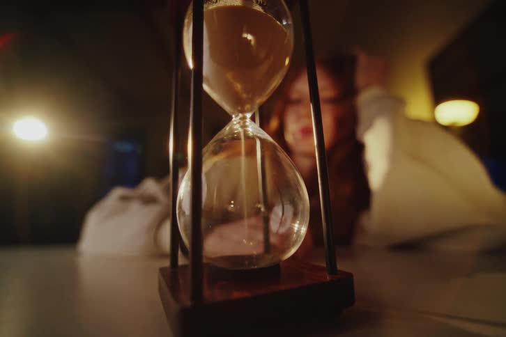 A woman turns over an hourglass sand timer in slow motion in a dark and moody room