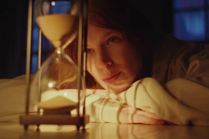 A thoughtful woman with red hair watches the sand fall in an hourglass in a warm, dimly lit room