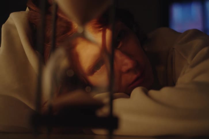 A pensive woman watches time slip away through an hourglass in a dimly lit, moody room
