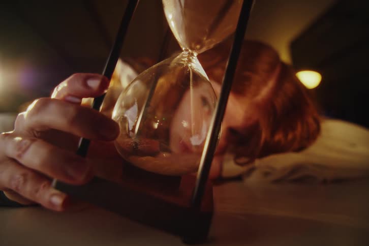 A contemplative woman lies down, her face distorted through the glass as she watches the sand pass through an hourglass in a dimly lit room