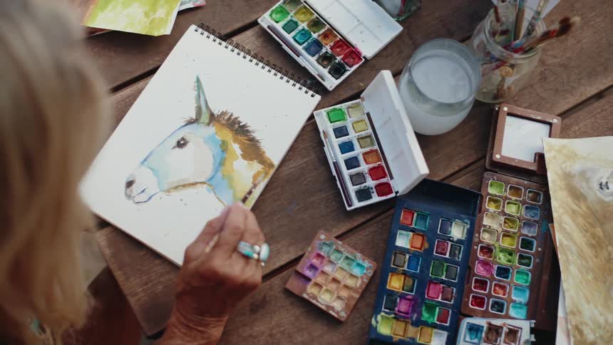 An elderly artist finds her creative flow while painting a colorful donkey with watercolors at an outdoor wooden table