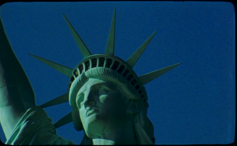 A vintage film close-up of the Statue of Liberty's face and crown against a deep blue sky, which transitions into a chaotic and blurry shot